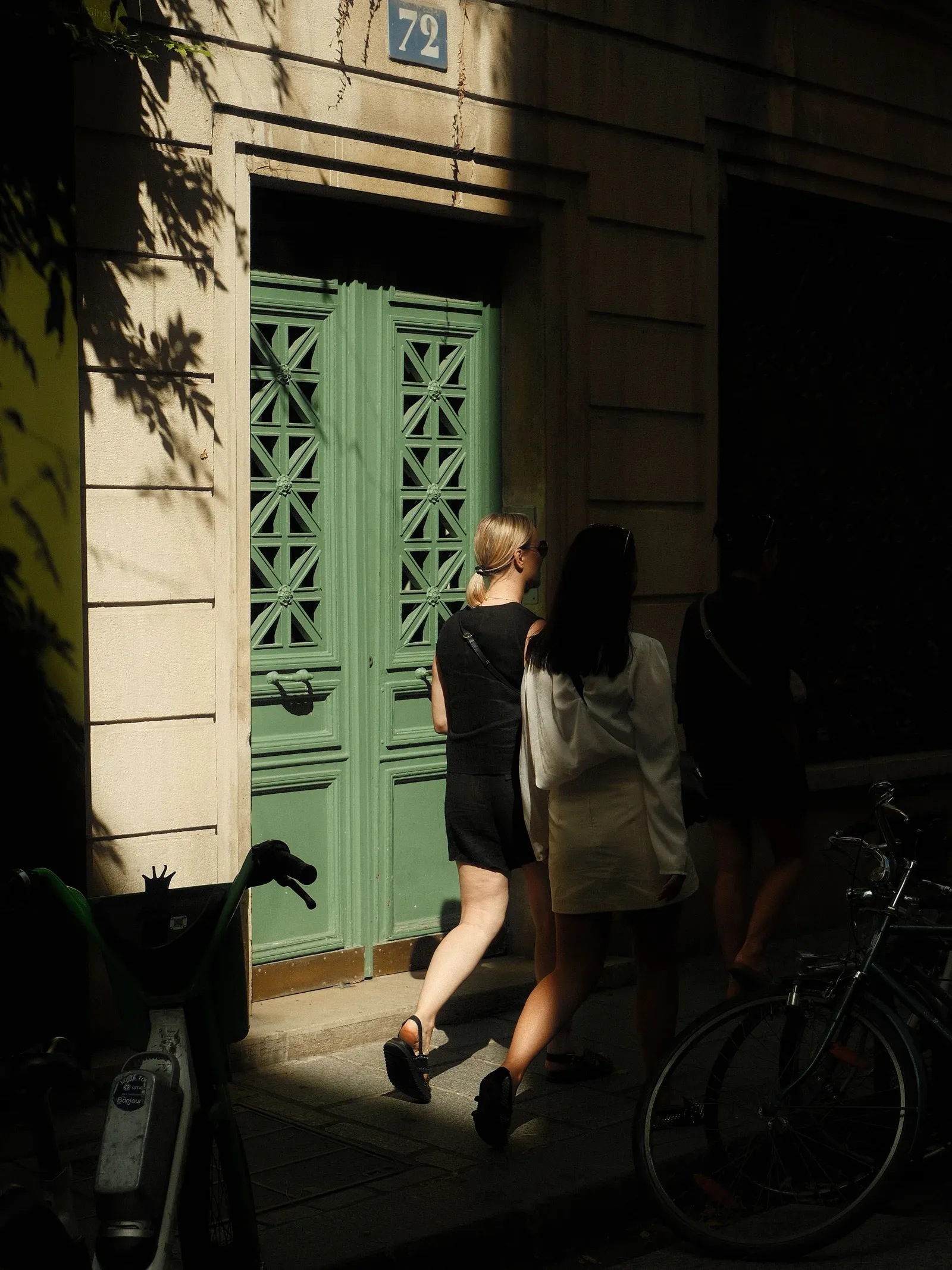 Two people walk past a tall green wooden door with carved lattice panels, in a sunlit stone building. Bicycles are parked in the shade.